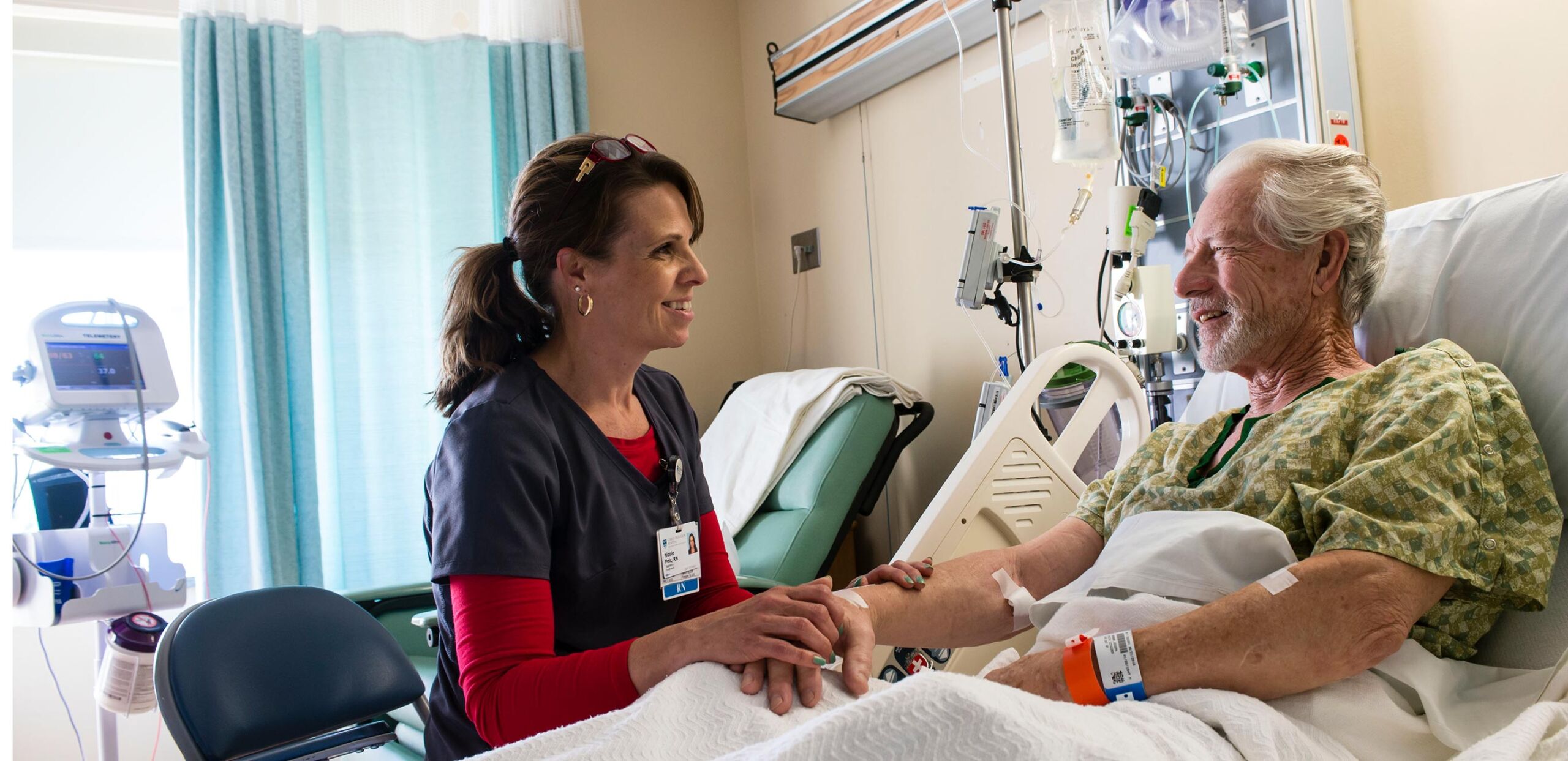 Friendly female nurse holds hand of smiling older male patient while they talk in his room at Cooley Dickinson Hospital, 30 Locust Street, Northampton, MA 01060.