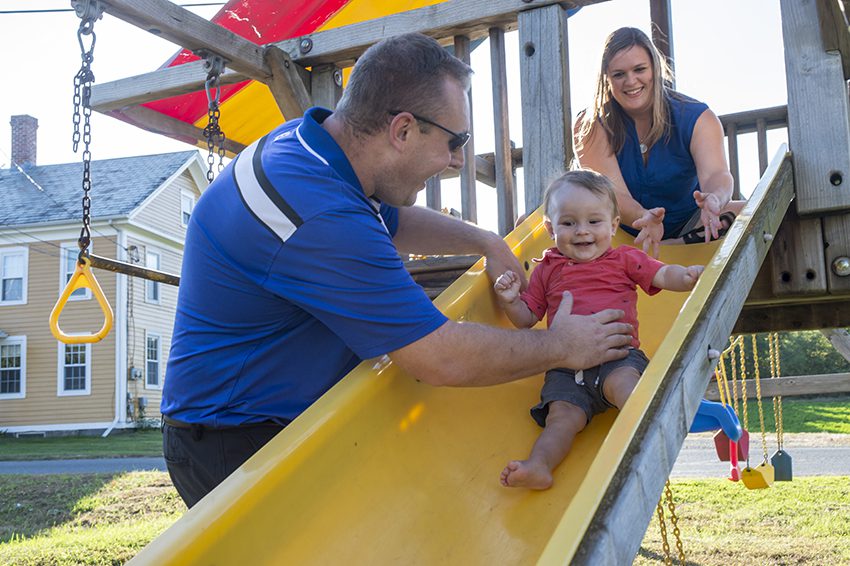 A man smiling and helping his baby down a playground slide while his wife watches from above