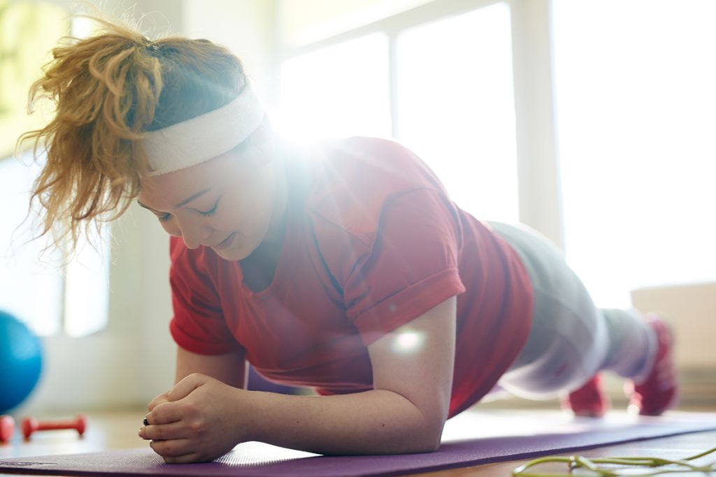 Portrait of young obese woman working out on yoga mat in fitness studio: holding plank exercise with effort to lose weight