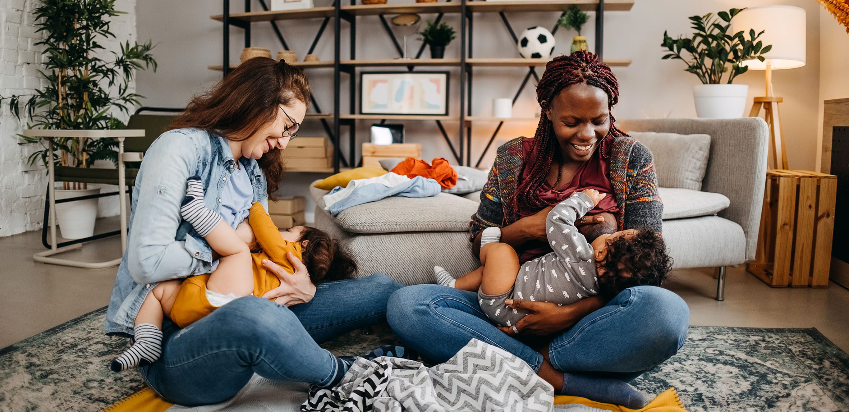 Two women sitting on the loving room floor breastfeeding their infants