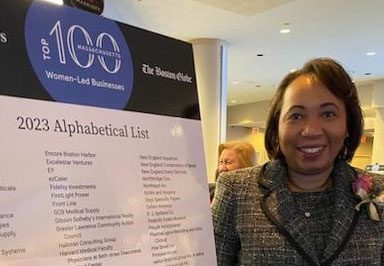 Lynnette Watkins standing next to a poster recognizing the top 100 women-led businesses in Massachusetts